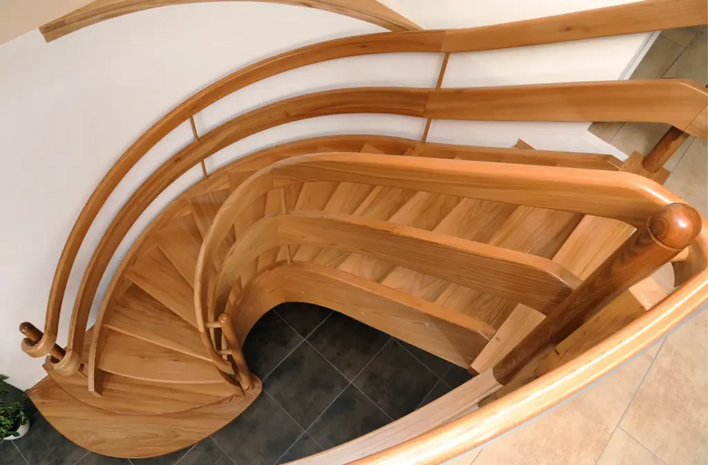 Blick von oben auf eine geschwungene Holztreppe mit glatten, polierten Handläufen und Stufen vor einem gefliesten Boden und einer weißen Wand.