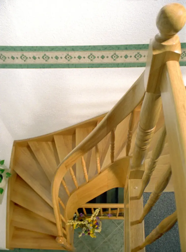 Draufsicht auf eine hölzerne Wendeltreppe mit einem dekorativen Geländer, die zu einem gefliesten Boden mit einem Blumenarrangement am unteren Ende führt; ein Teil einer grün gemusterten Wandeinfassung ist sichtbar.