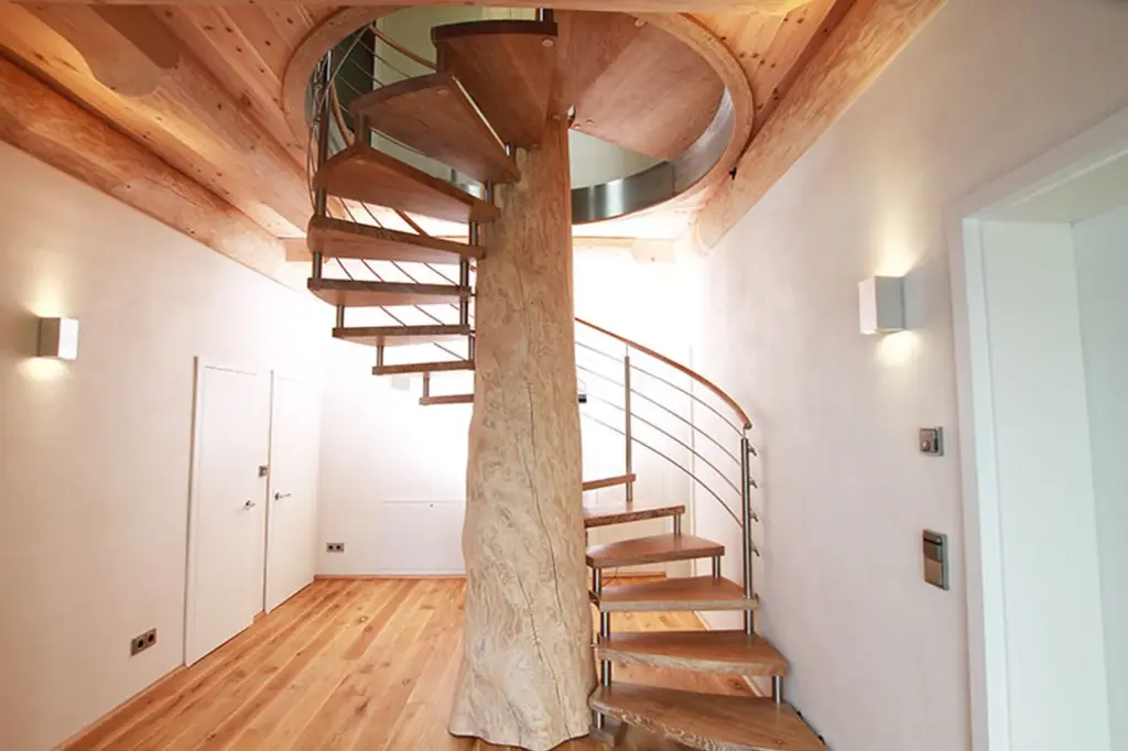 Eine moderne Spindeltreppe mit Holzstufen und einer zentralen Holzsäule steht in einem hellen Raum mit hellen Wänden und Holzböden. Die Treppe hat ein elegantes Metallgeländer.