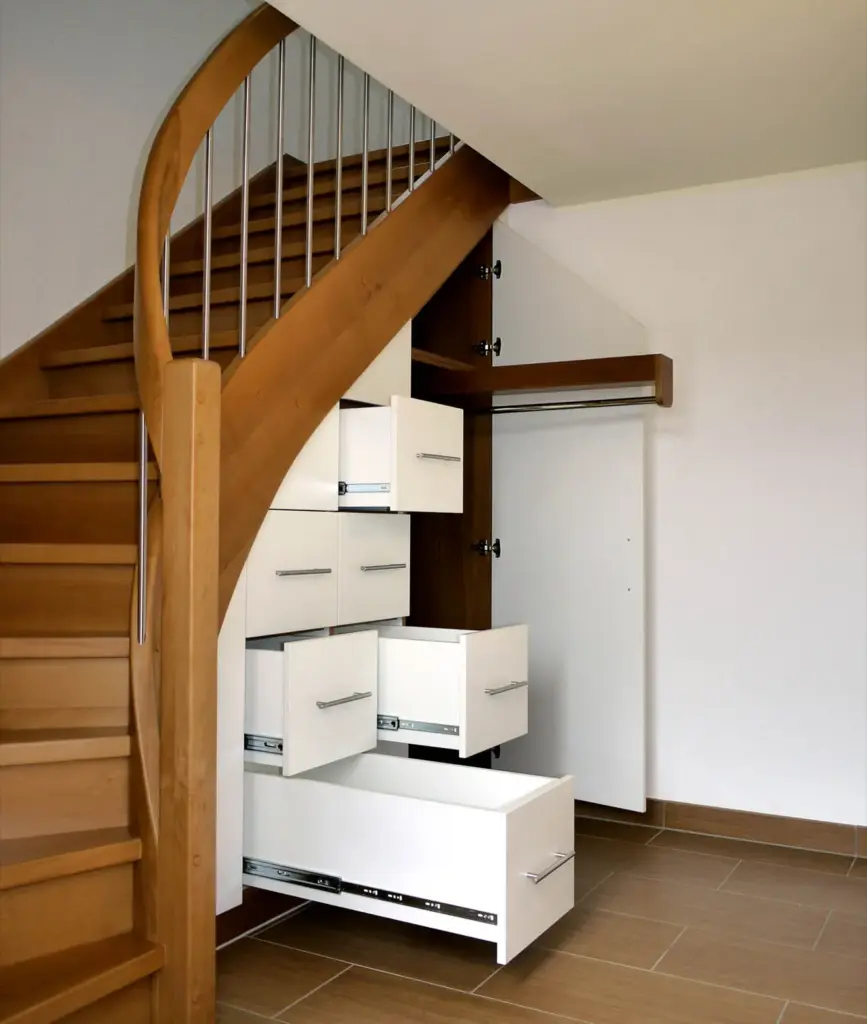Ein Satz weißer Schubladen, die unter einer Holztreppe installiert sind und den Raum in einem modernen Interieur effizient als Stauraum nutzen. Die Treppe hat ein Metallgeländer und einen braunen Fliesenbelag.