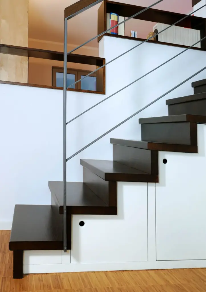 Eine moderne Treppe mit dunklen Holzstufen und einem eleganten Metallgeländer, gebaut über weißen Schränken mit runden Griffen, auf einem hellen Holzboden in einem zeitgenössischen Wohnbereich.