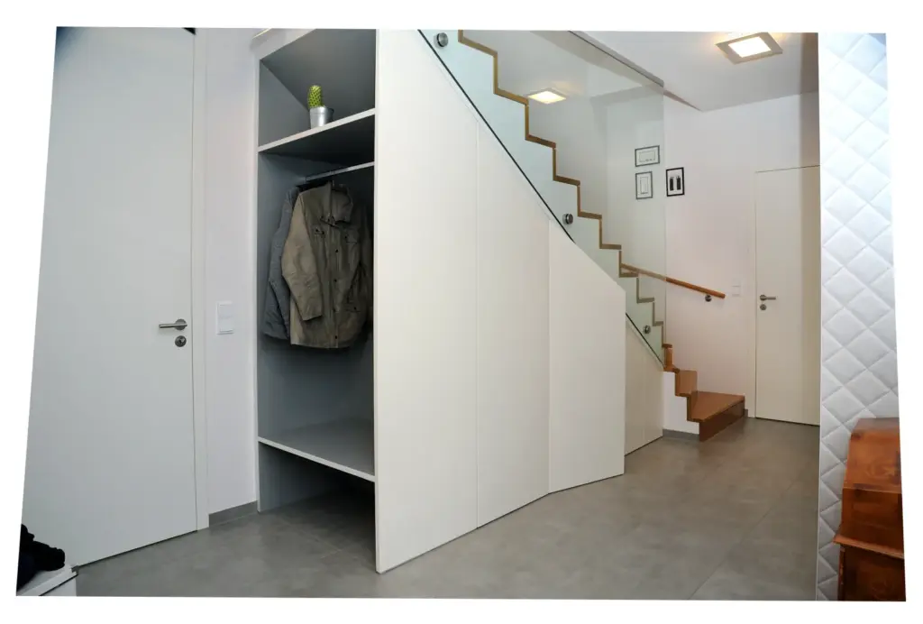 Ein moderner Flur mit einer Holztreppe, einem eingebauten Stauraum unter der Treppe für Mäntel und einem Regal mit einer kleinen Pflanze. Der Raum hat weiße Wände, geflieste Böden und eine minimalistische Einrichtung.