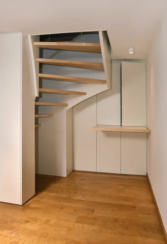 Eine moderne, minimalistische Holztreppe mit offenen Setzstufen und weißen Stützen in einem kleinen Raum mit hellem Holzfußboden und weißen Wänden. Unter der Treppe befinden sich ein eingebautes Regal und Schränke.