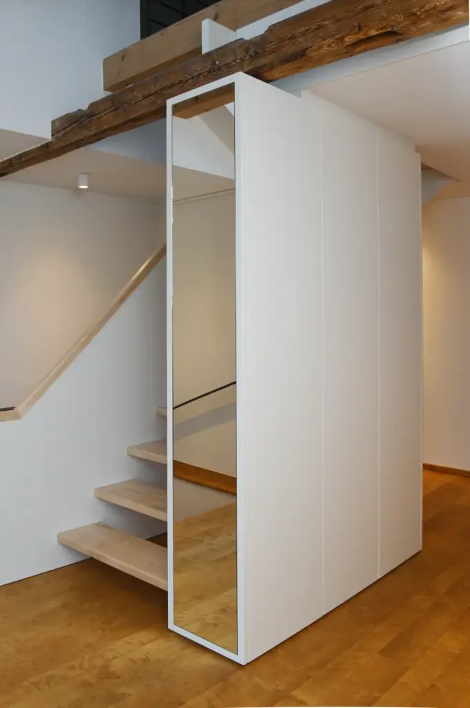 Ein modernes Interieur mit einer schwebenden Holztreppe neben einem großen, minimalistischen weißen Kleiderschrank mit einem Ganzkörperspiegel an einer Seite. Holzböden und freiliegende Deckenbalken verleihen dem Raum ein modernes Aussehen.