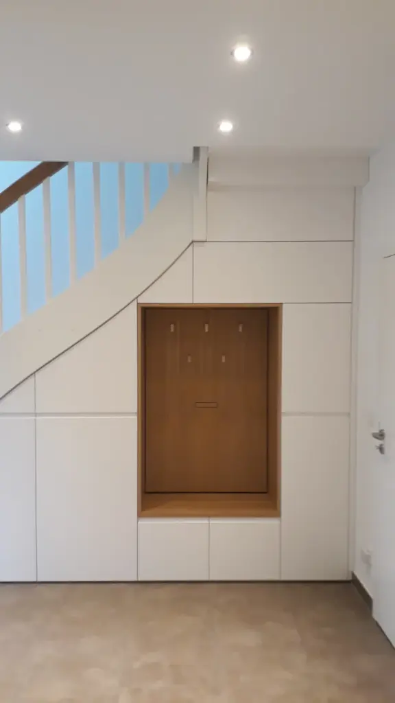 Ein moderner, minimalistischer Flur mit weißen Einbauschränken unter einer Treppe, mit einer eingelassenen Holzecke und Kleiderhaken in der Mitte. Der Raum ist durch Deckeneinbauleuchten hell erleuchtet.