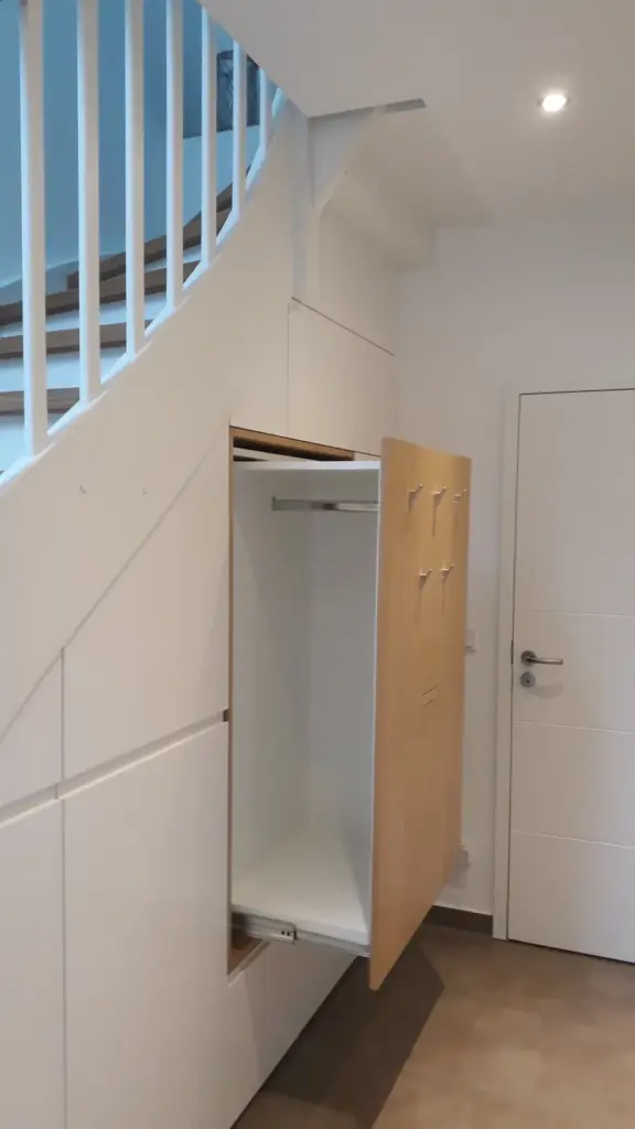 Ein moderner Flur mit weißen Wänden und einer Treppe. Unter der Treppe befindet sich ein ausziehbarer Einbauschrank mit Haken an der Tür und einer Kleiderstange im Inneren. In der Nähe befindet sich eine weiße Tür mit einem silbernen Griff.