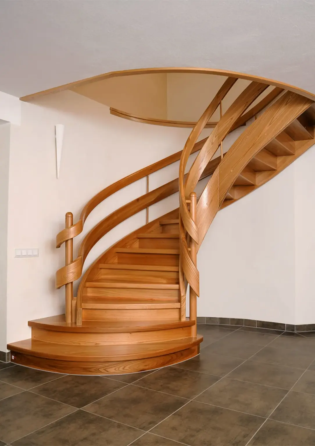 Eine moderne Holzspindeltreppe mit geschwungenem Geländer und polierten Stufen vor einer weißen Wand und einem gefliesten Boden in einem modernen Innenraum.