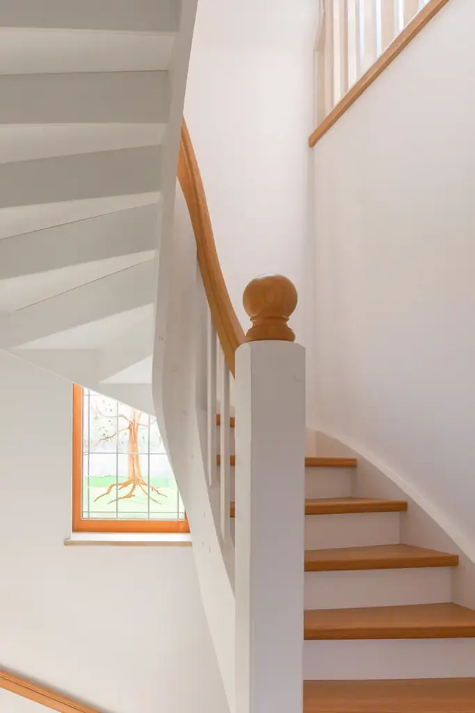 Eine Holztreppe mit weißem Geländer und einem polierten Holzhandlauf windet sich neben einer Wand nach oben. Links ist ein Buntglasfenster mit einem Baummotiv zu sehen. Die Szene ist gut beleuchtet und minimalistisch.