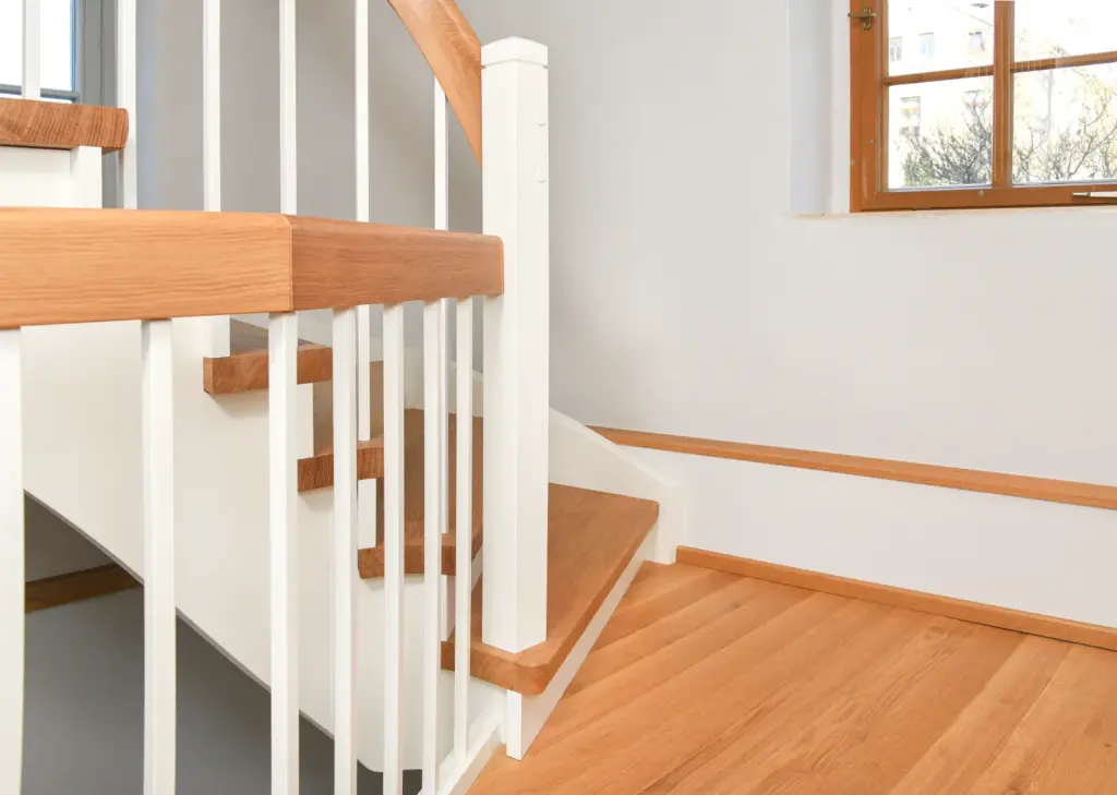 Eine moderne Holztreppe mit weißem Geländer und hellbraunen Stufen, neben einem Fenster, das natürliches Licht hereinlässt. Der Boden ist aus passendem hellem Holz gefertigt.