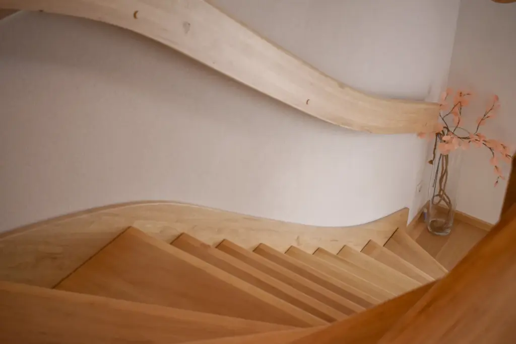 Eine spiralförmige Holztreppe mit glattem, geschwungenem Geländer führt an einer weißen Wand hinunter. In der unteren Ecke steht eine Vase mit hellrosa Kunstblumen.