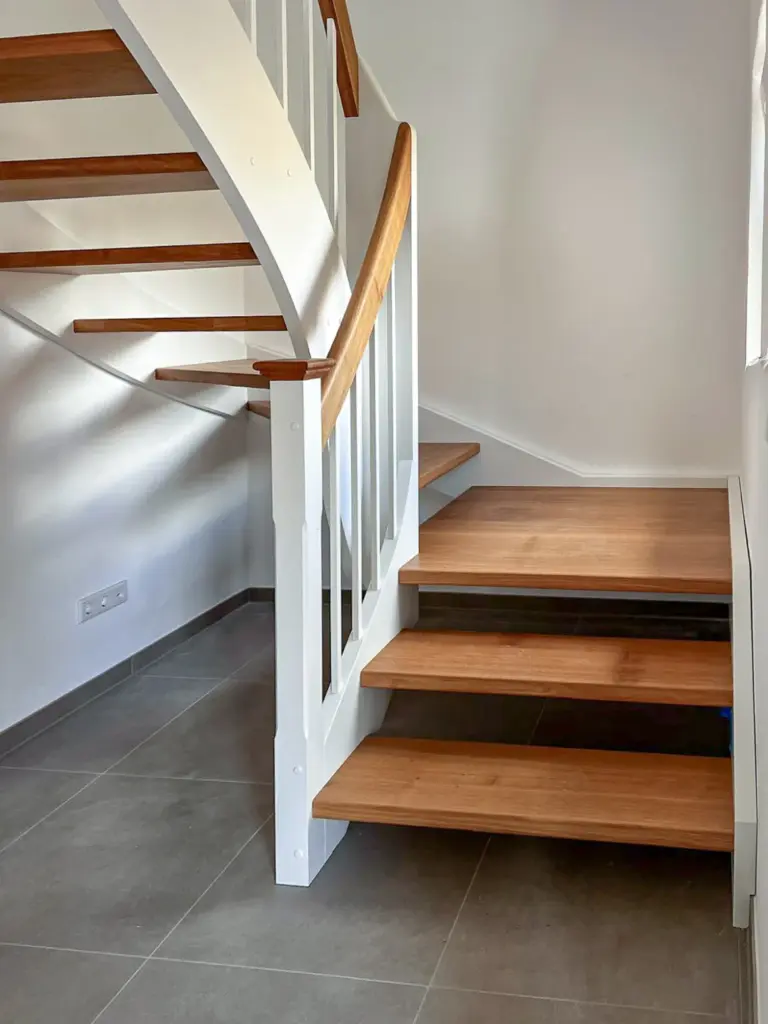Eine moderne Holztreppe mit weißem Geländer und braunen Stufen wird in einem hellen Raum mit grauem Fliesenboden und schlichten weißen Wänden gezeigt.