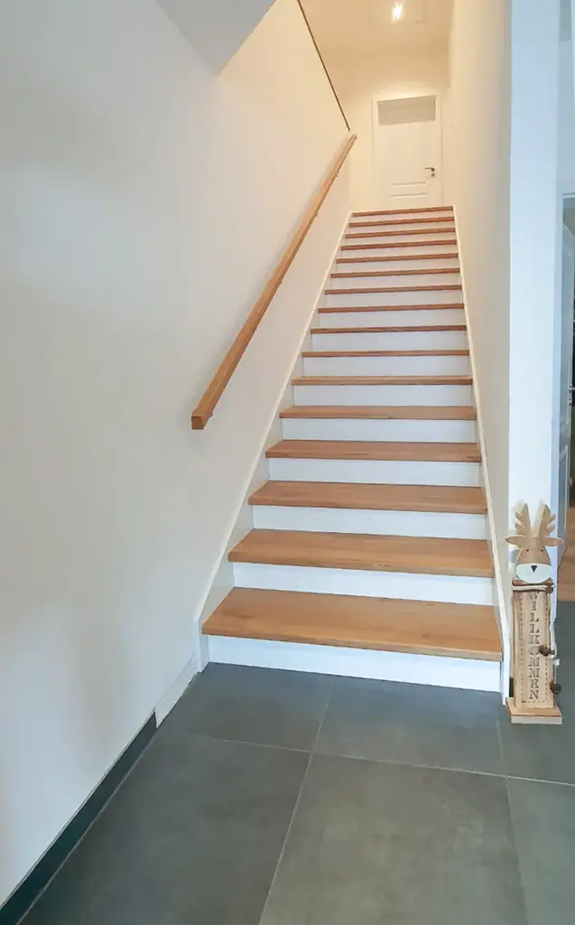 Eine moderne Innentreppe mit Holzstufen und einem einfachen Handlauf führt zu einer weißen Tür. Unten rechts steht eine kleine dekorative Rentierfigur auf dem Boden in der Nähe der ersten Stufe.