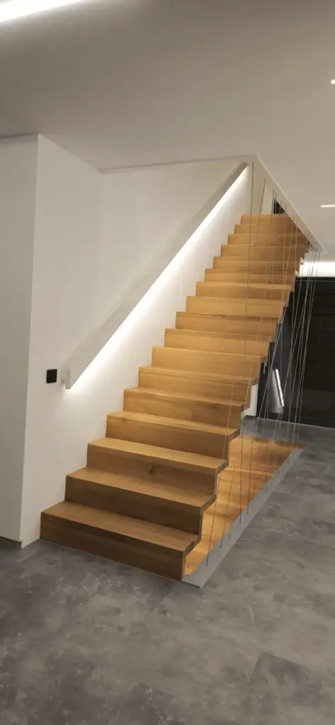 Eine moderne Innentreppe mit Holzstufen, weißen Seitenwänden, minimalistischem Drahtgeländer und eingebauter LED-Beleuchtung entlang der Wand, vor einer weißen Wand und grauem Fliesenboden.