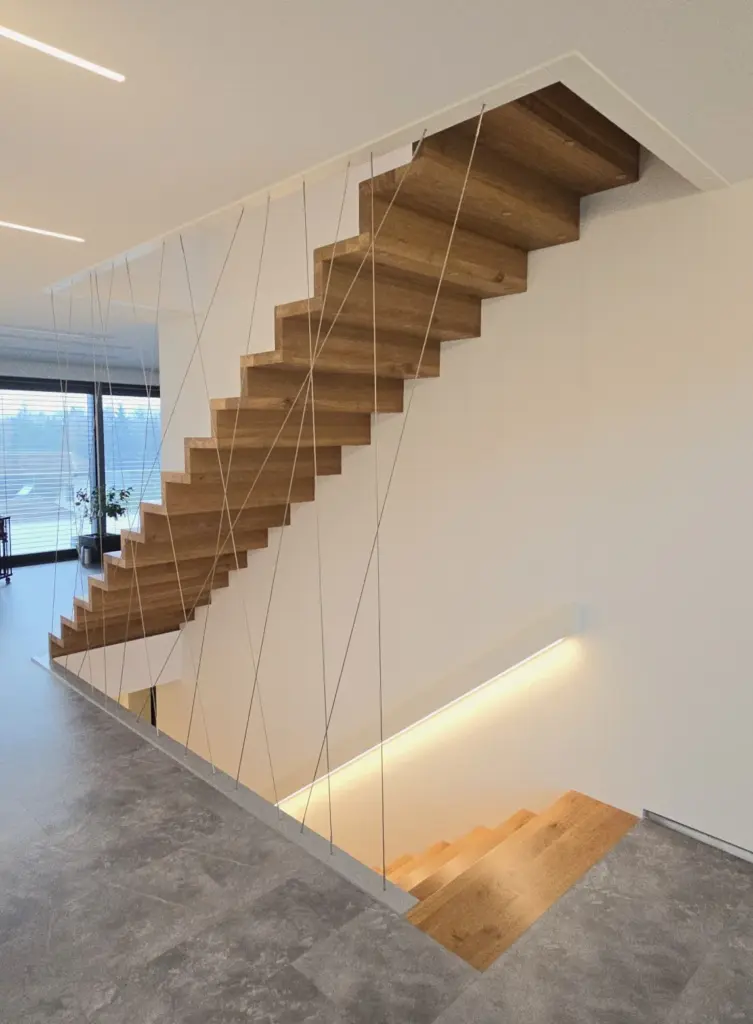 Eine moderne schwebende Holztreppe mit dünnen weißen Seilen als Stütze, die zwei Etagen in einem minimalistischen, hellen Innenraum mit grauem Fliesenboden und einem großen Fenster im Hintergrund verbindet.