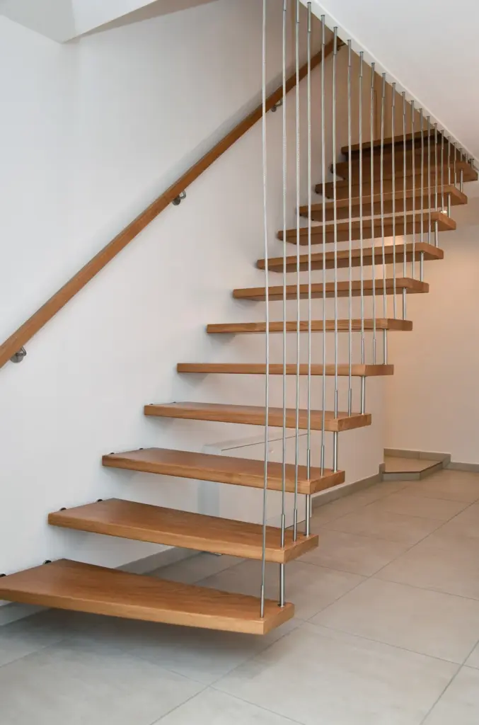 Moderne schwebende Holztreppe mit Metallseilen als Stütze, befestigt an einer weißen Wand in einem minimalistischen Innenraum mit Fliesenboden.