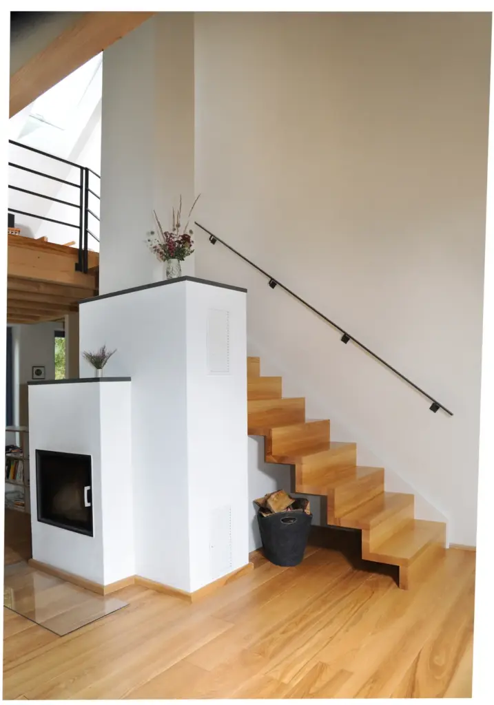 Moderne Holztreppe mit schwarzem Handlauf an einer weißen Wand, über einem weißen Kamineinsatz. Ein Korb mit Brennholz steht auf dem Boden, und eine Vase mit Blumen steht auf dem Kamin.