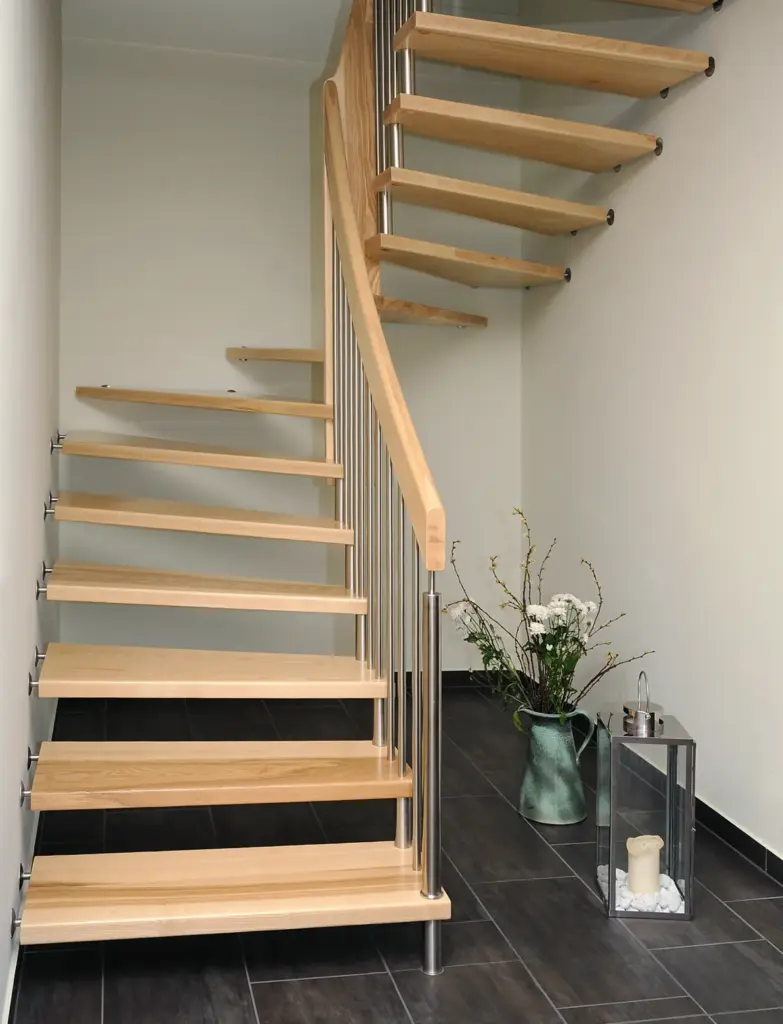 Eine moderne Holztreppe mit offenen Setzstufen und Metallgeländer schlängelt sich in einem minimalistischen Flur nach oben. Auf dem dunklen Kachelboden unter der Treppe stehen eine Vase mit weißen Blumen und eine große Laterne mit einer Kerze.