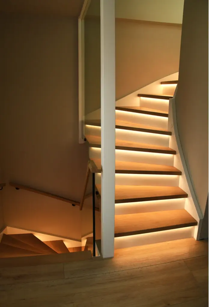 Eine moderne Treppe mit Holzstufen wird von einer warmen Einbauleuchte unter jeder Stufe beleuchtet. Die Treppe biegt an einem Podest ab und führt weiter nach oben, begrenzt durch schlichte weiße Wände und Glasgeländer.