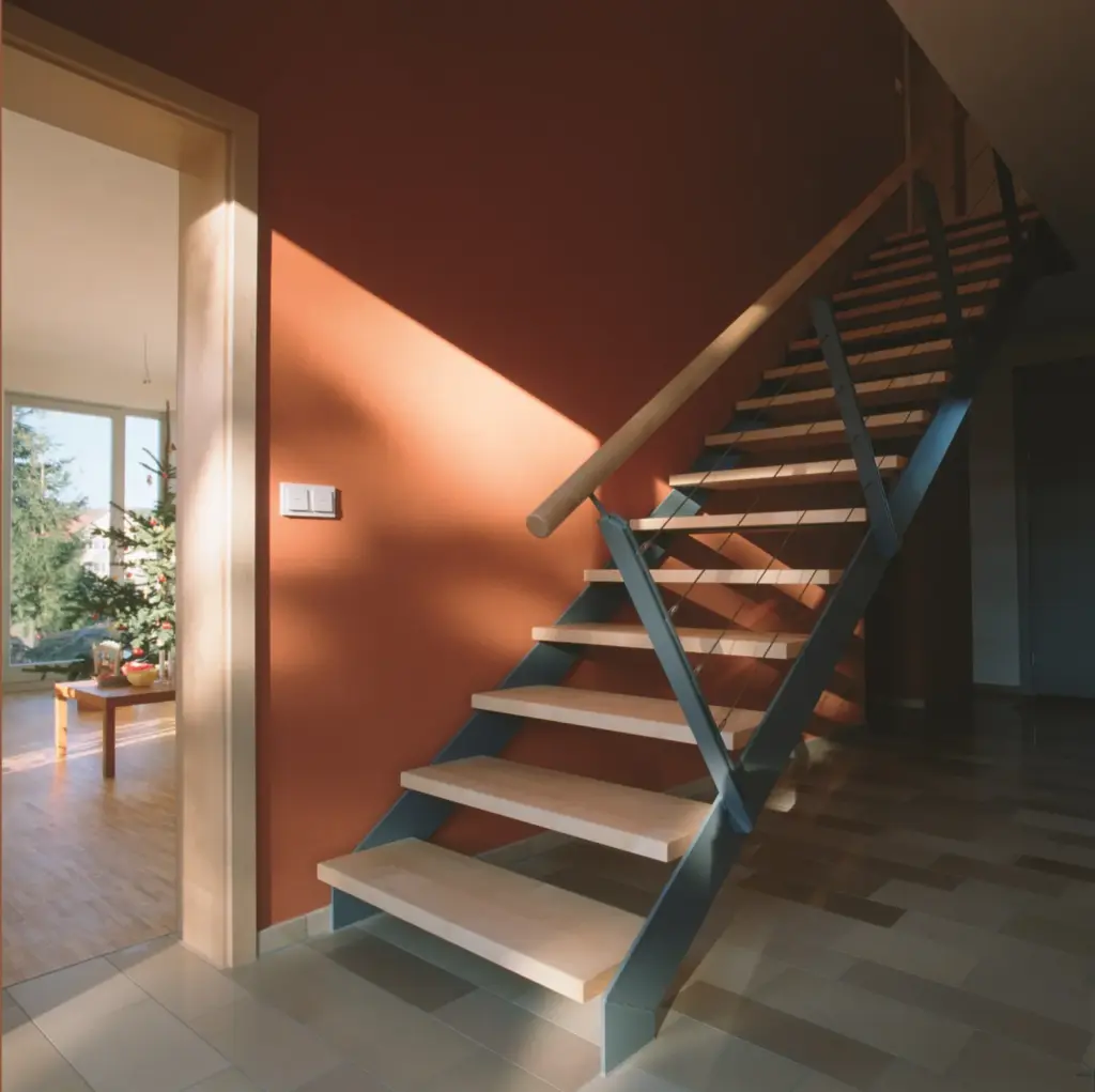 Ein modernes Interieur mit einer offenen Holztreppe und einem Metallgeländer vor einer rötlich-braunen Wand. Sonnenlicht fällt ein und wirft Schatten auf den gefliesten Boden. Im angrenzenden Raum ist ein geschmückter Weihnachtsbaum zu sehen.