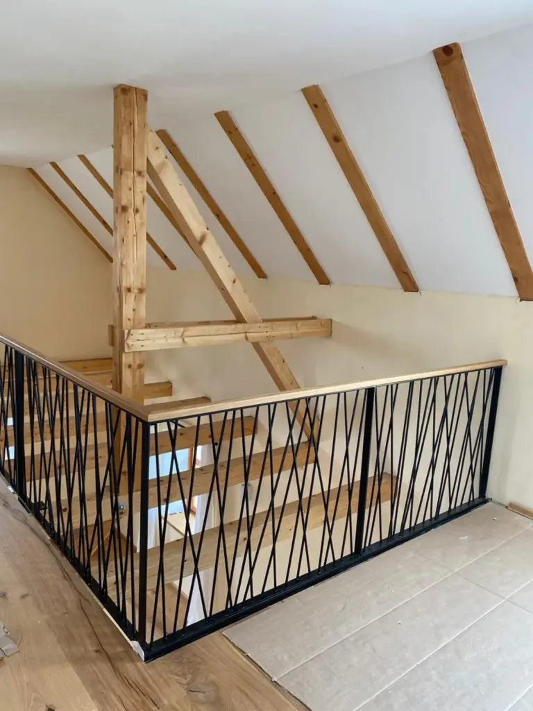 Eine Holztreppe mit schwarzem Metallgeländer führt zu einem oberen Dachraum mit schräger weißer Decke und freiliegenden Holzbalken. Der Boden ist aus Holz und mit Kartonplatten ausgelegt.