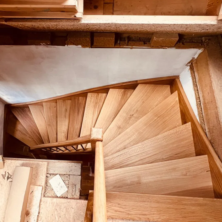Draufsicht auf eine hölzerne Wendeltreppe mit unvollendeten Baumaterialien und einer freiliegenden Wand um sie herum. Die Treppe ist sauber eingebaut, aber die Umgebung wirkt unvollständig.