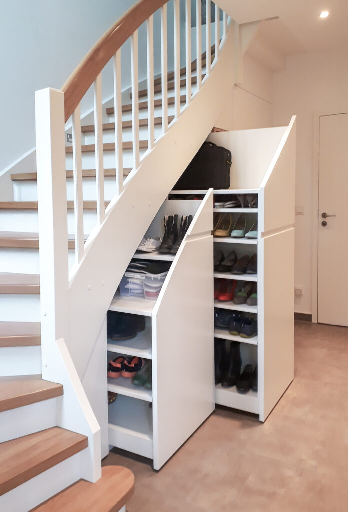 Weiße Treppe mit Holzstufen und einem geschwungenen Geländer. Unter der Treppe befinden sich zwei ausziehbare Schränke mit Schuhen, Aufbewahrungsboxen und einer schwarzen Tasche obenauf. Der Bereich ist aufgeräumt und modern.
