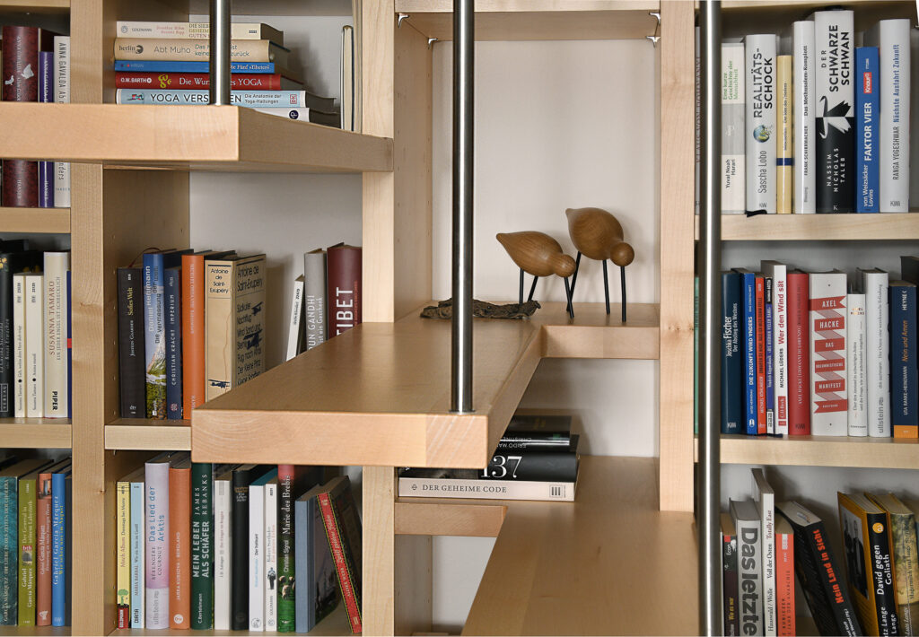 Bücherregale, gefüllt mit verschiedenen Büchern in deutscher und englischer Sprache. Ein Paar hölzerne Vogelfiguren sind auf einem Regal in der Mitte ausgestellt. Die Regale sind aus hellem Holz mit Metallstützen und einem offenen, modernen Design.
