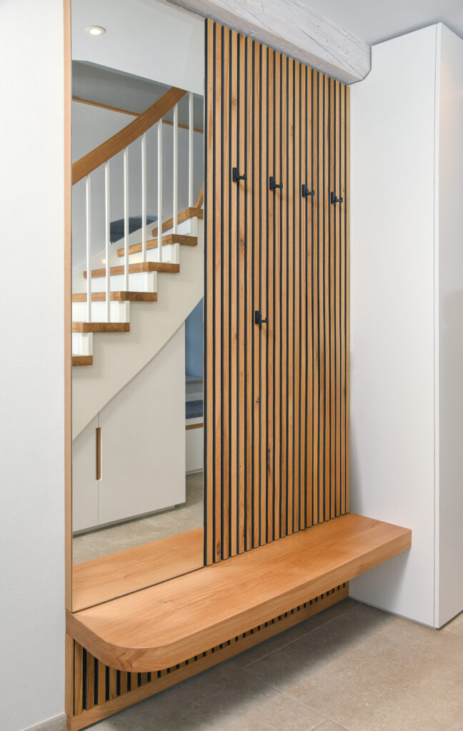 Moderner Eingangsbereich mit einer Holzlattenwand, sechs schwarzen Haken, einem großen Spiegel und einer eingebauten Holzbank. Eine Treppe mit weißem Geländer und Holzstufen spiegelt sich im Spiegel.