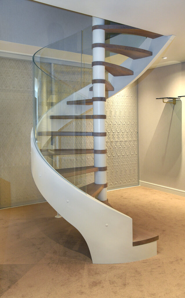 Eine moderne Wendeltreppe mit hölzernen Stufen, einer weißen Mittelsäule und einem Glasgeländer steht in einem Raum mit Teppichboden, gemusterten Tapeten und gedämpfter Beleuchtung.