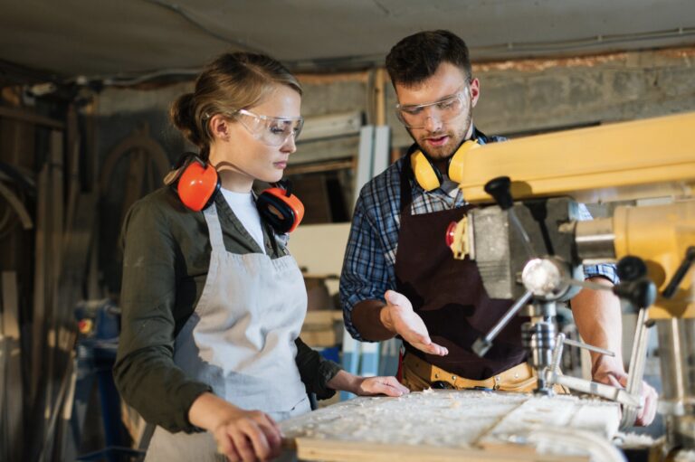 Zwei Personen mit Schutzbrillen und Schürzen arbeiten in einer Holzwerkstatt. Eine Person zeigt auf eine Maschine, während sie etwas erklärt, und die andere hört mit einem Gehörschutz im Nacken aufmerksam zu.