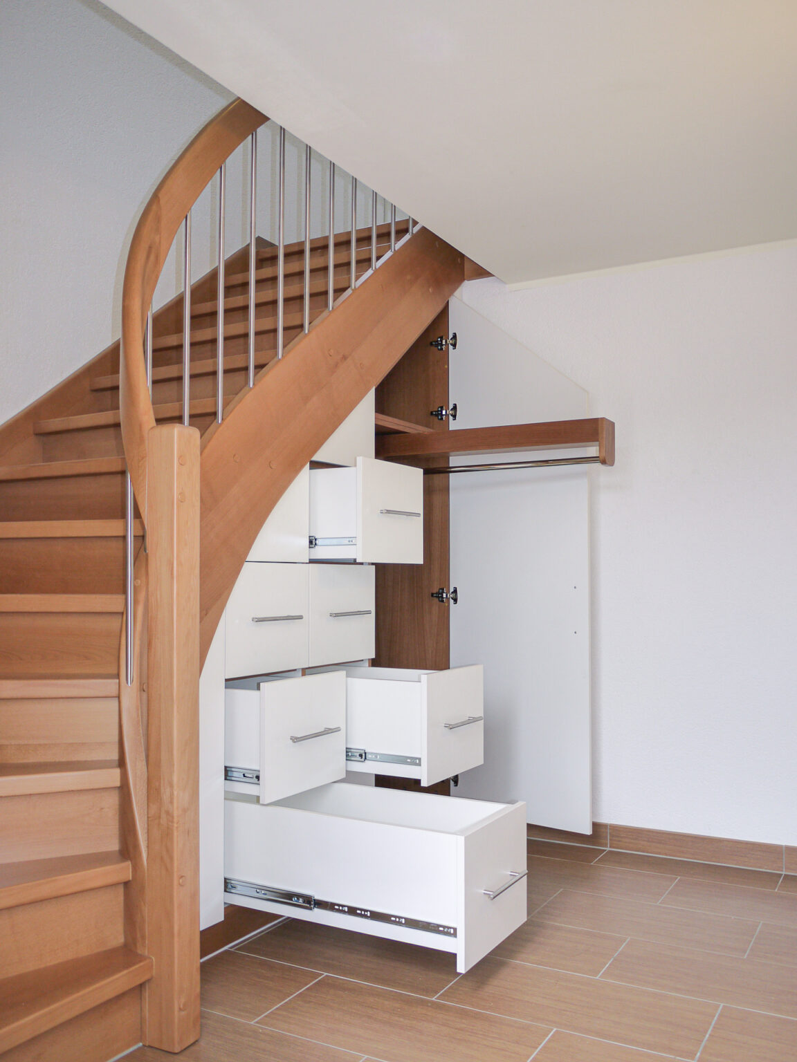 Eine Holztreppe mit eingebauten weißen Schubladen und Schränken darunter, von denen einige offen sind, um den Platz optimal zu nutzen. Der Boden ist hellbraun gefliest, die Wände sind weiß.