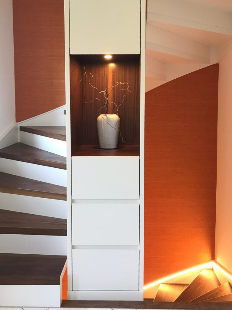 Eine moderne Treppe mit Holzstufen und weißen Setzstufen, die mit orangefarbenen Wandpaneelen versehen ist. In einer eingebauten Nische mit einem Strahler steht eine weiße Vase mit dekorativen Zweigen. Die unteren Stufen werden durch eine warme Beleuchtung hervorgehoben.