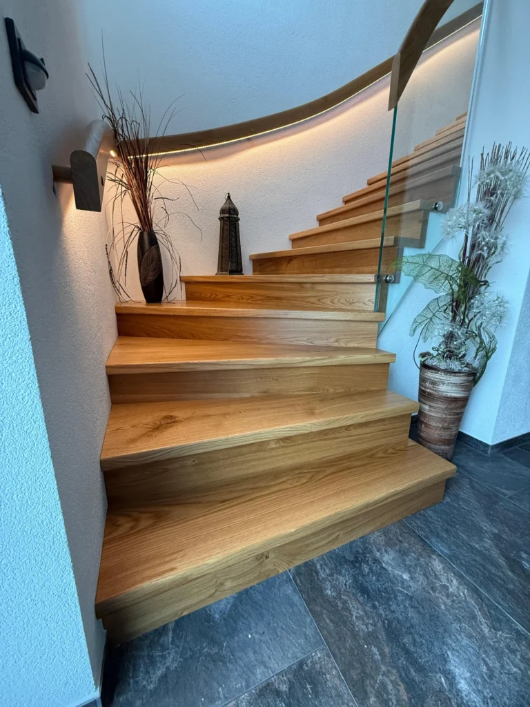 Eine moderne Holztreppe mit Glasgeländer schlängelt sich nach oben, dekoriert mit Topfpflanzen, hohen Vasen und sanfter Wandbeleuchtung auf einem grauen Fliesenboden.