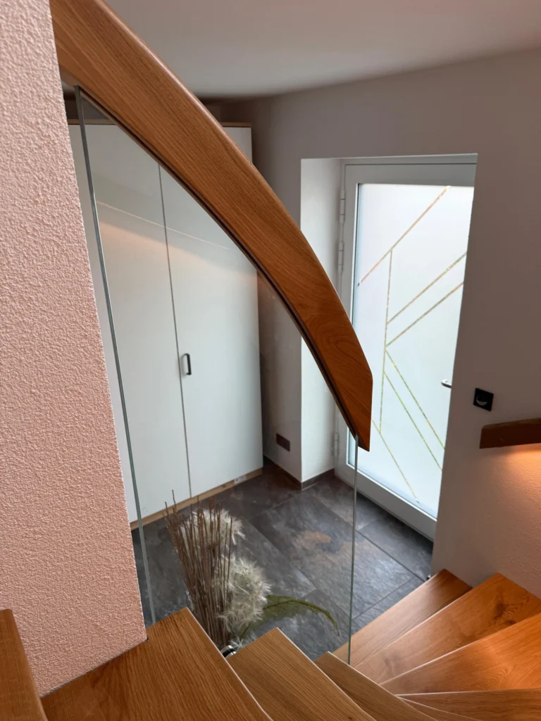 Blick von einer Holztreppe hinunter in einen modernen Eingangsbereich mit einer Glastür, einem weißen Schrank, einem dunklen Fliesenboden und einer Vase mit getrockneten Pflanzen in der Nähe der Ecke.