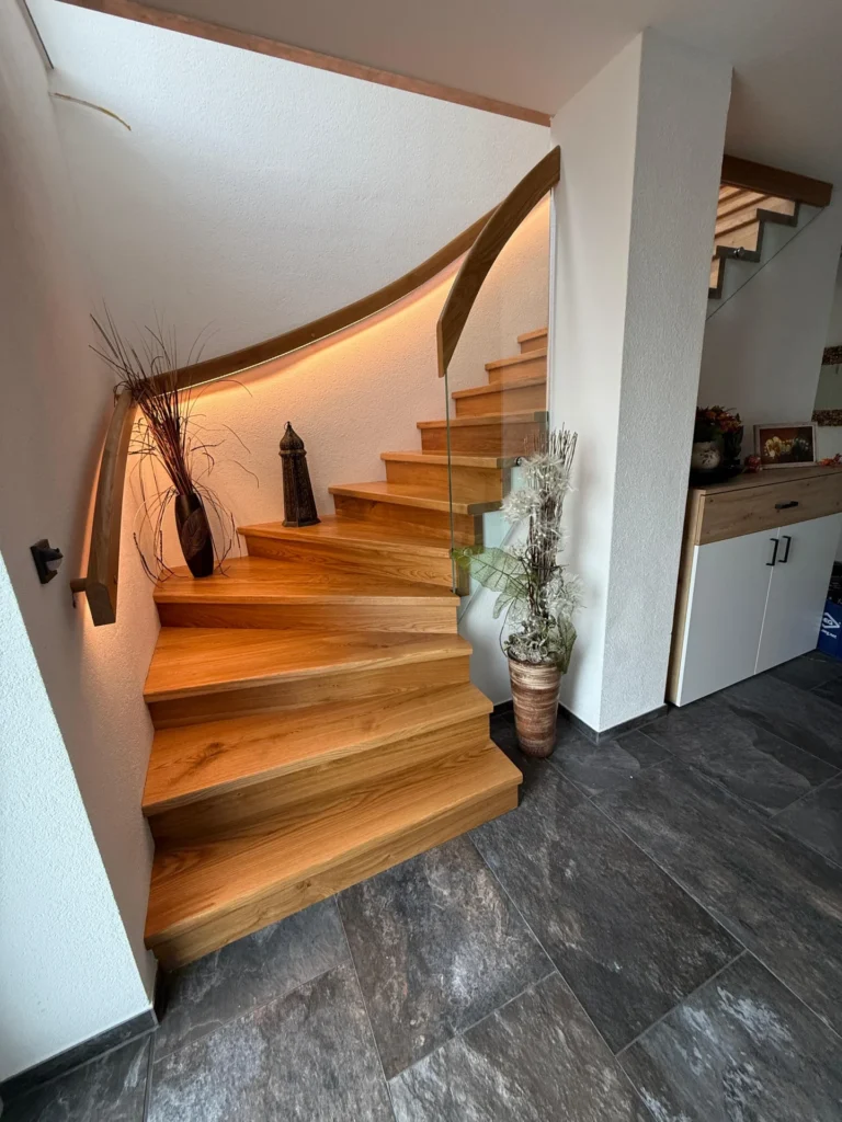 Eine moderne Holztreppe mit einem Glasgeländer schlängelt sich nach oben, akzentuiert durch warme Beleuchtung. Auf den Stufen und dem Treppenabsatz stehen dekorative Vasen mit getrockneten Pflanzen. Der Boden ist mit großen grauen Fliesen ausgelegt.