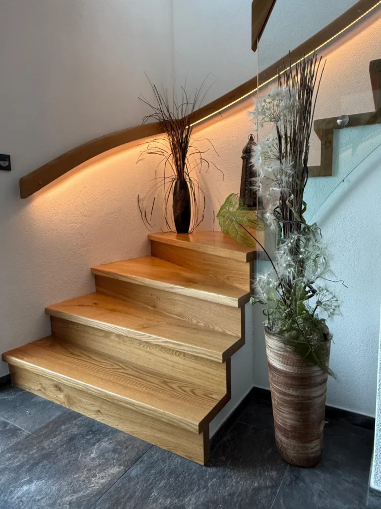 Eine moderne Holztreppe mit drei Stufen, dekoriert mit hohen Vasen mit getrockneten Pflanzen, steht vor einer weißen Wand mit einem geschwungenen Holzhandlauf und einer weichen, warmen Akzentbeleuchtung. Der Boden besteht aus dunklen Steinfliesen.