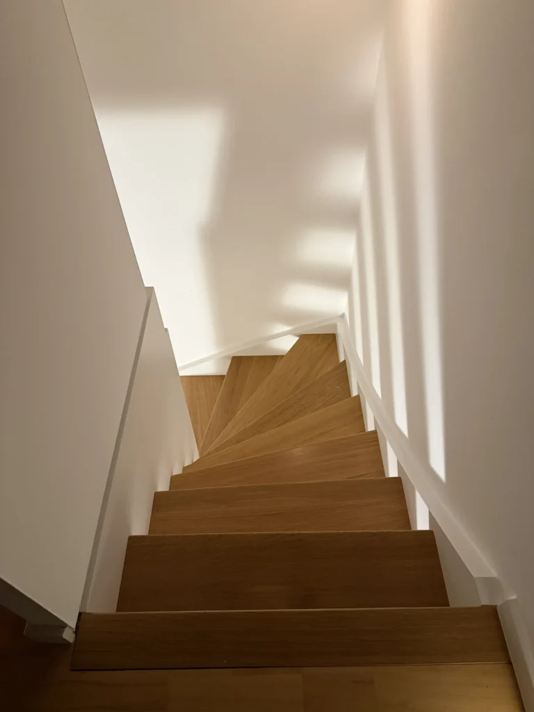 Draufsicht auf eine schmale hölzerne Wendeltreppe mit weißen Wänden und Sonnenlicht, das geometrische Schatten an die Wand wirft.