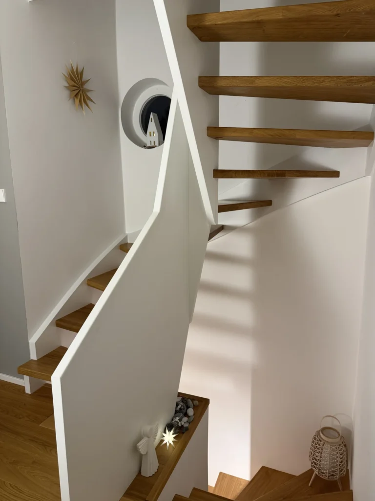 Eine moderne Holztreppe mit offenen Setzstufen und weißem Geländer, mit minimalistischem Dekor, darunter ein Stern an der Wand, ein kleines Hausornament in einem runden Fenster und eine dekorative Laterne auf dem Boden.
