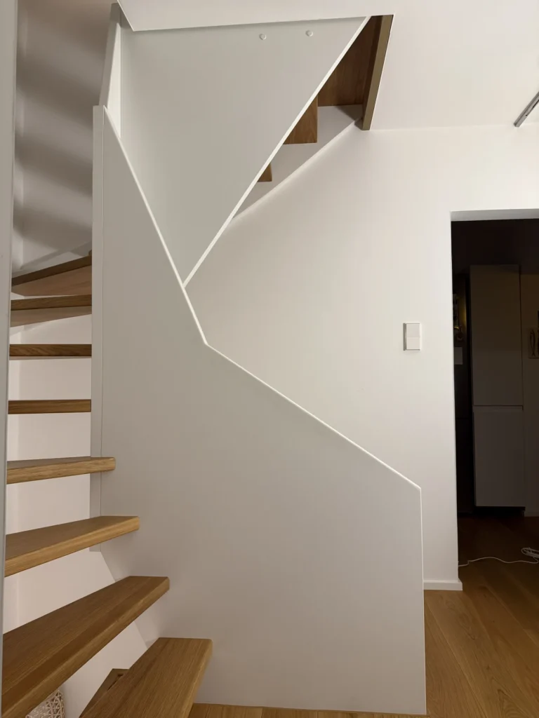 Eine moderne Holztreppe mit einem kantigen, geometrischen weißen Geländer, offenen Setzstufen und klaren Linien führt in einem minimalistischen Interieur mit hellem Holzboden und weißen Wänden nach oben.