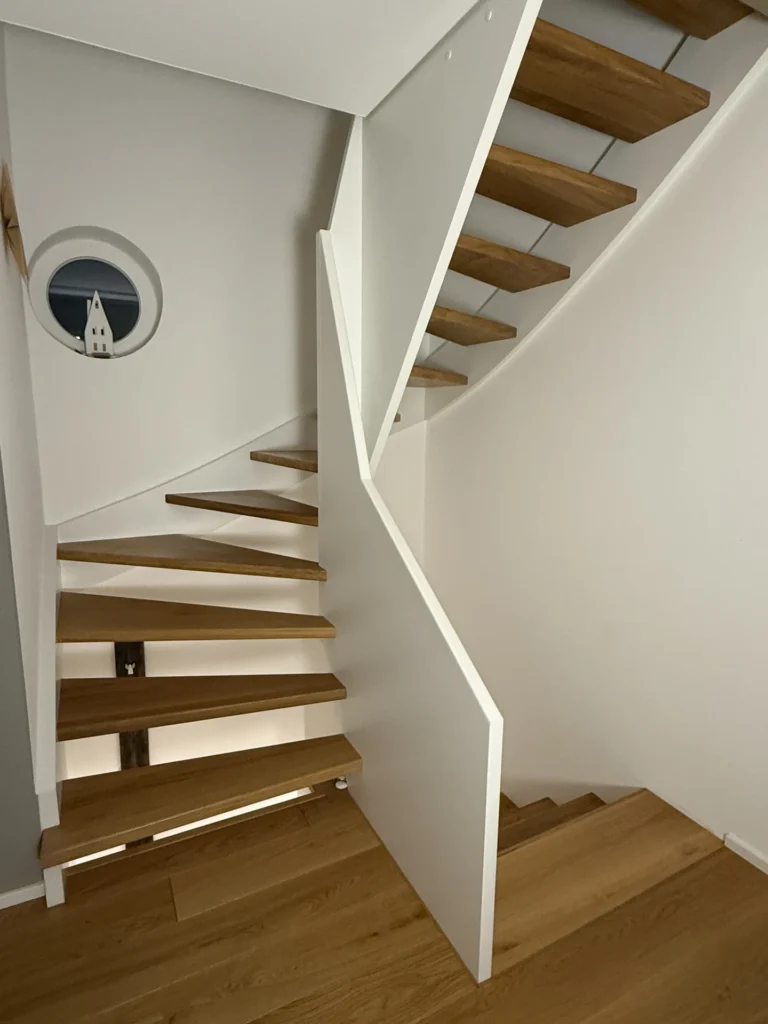 Eine moderne Holztreppe mit offenen Setzstufen windet sich nach oben, umschlossen von weißen Wänden. Ein kleines rundes Fenster auf der linken Seite zeigt den Blick auf einen entfernten Kirchturm vor einem dunklen Himmel.