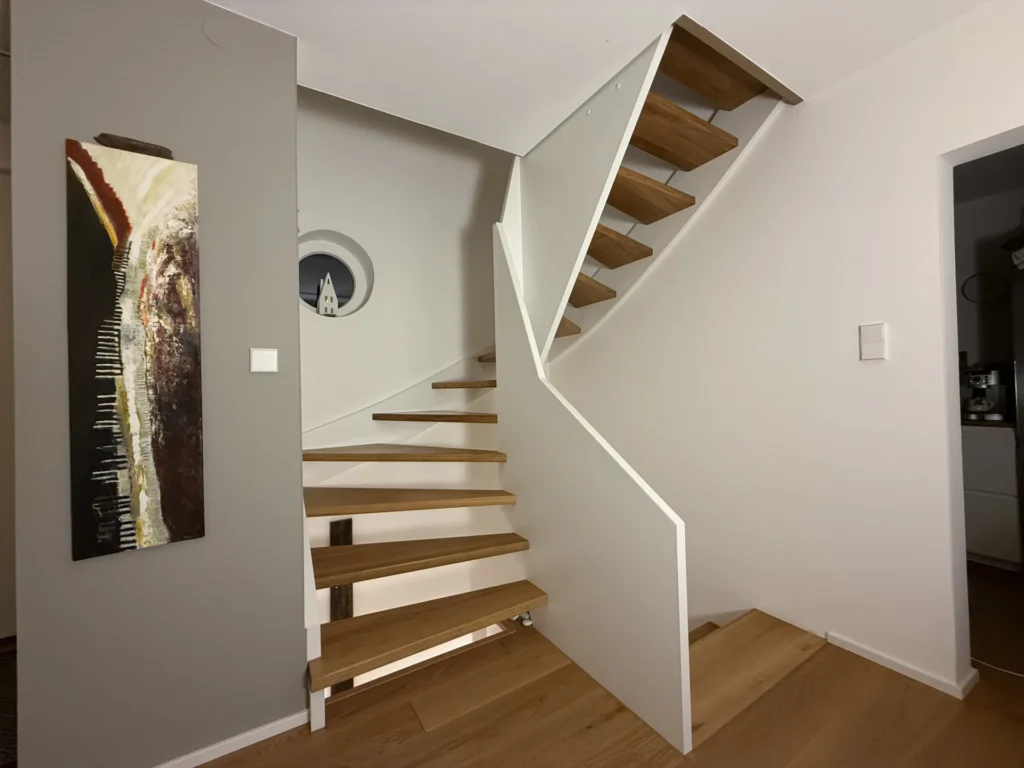 Eine moderne Holztreppe mit offenen Setzstufen und weißen Seitenwangen führt in zwei Richtungen in einem modernen Haus nach oben. Neben der Treppe sind abstrakte Wandkunst und ein rundes Fenster zu sehen.