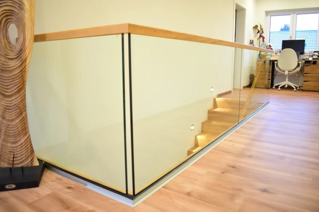 Eine moderne Holztreppe mit Glasgeländerpaneelen und einem Holzhandlauf führt zu einem Büroraum mit Schreibtisch und Stuhl in der Nähe eines Fensters. Der Boden ist aus hellem Holz.