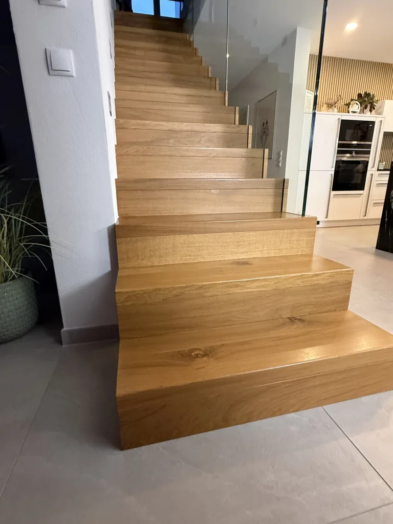 Eine moderne Holztreppe mit breiten Stufen und Glasgeländer, die entlang einer weißen Wand in einem zeitgenössischen Wohnbereich nach oben führt. Eine Küche und eine Topfpflanze sind teilweise im Hintergrund zu sehen.