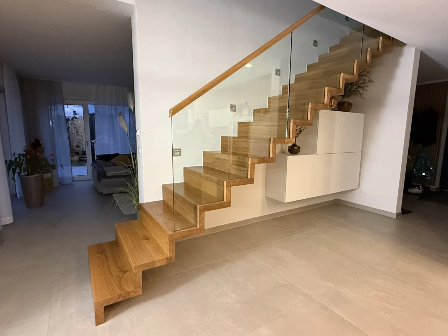 Moderne Holztreppe mit Glasgeländer und schwimmenden Stufen, die an einer weißen Wand hinaufführt. Minimalistisches Dekor mit Regalen für Vasen und Pflanzen. Der darunter liegende Bereich ist offen und schafft ein geräumiges und stilvolles Interieur.