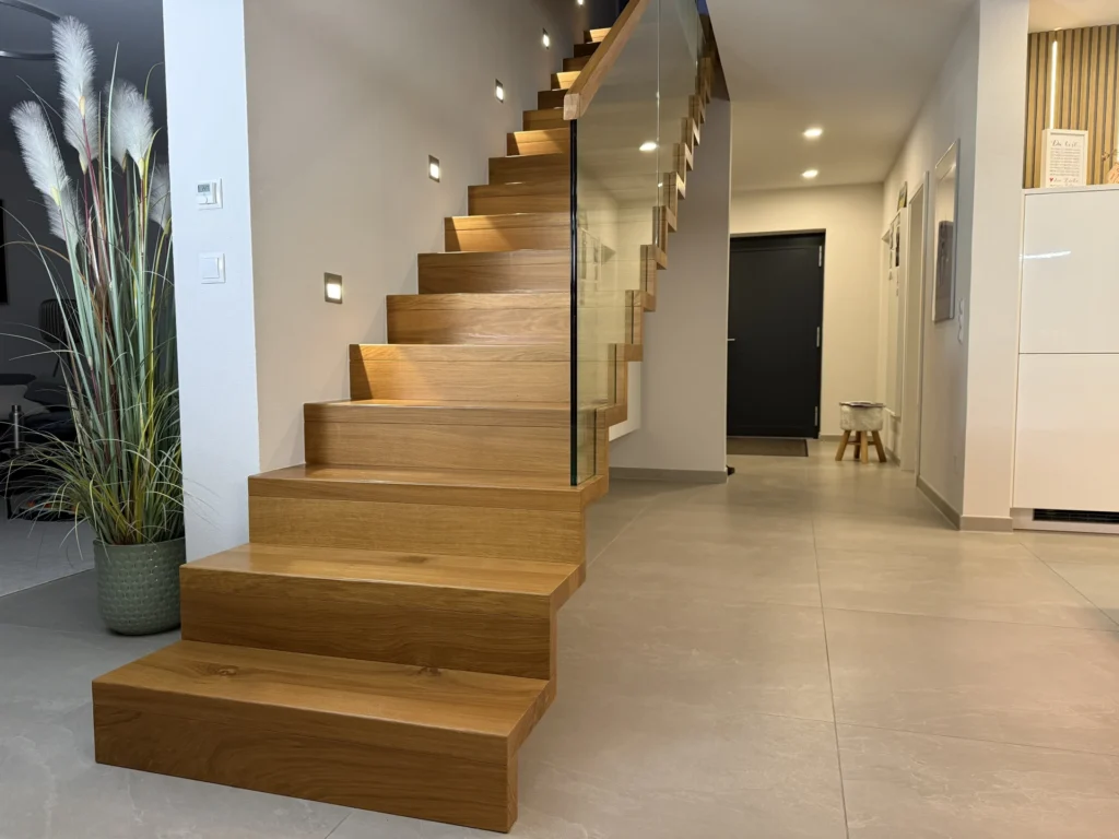 Ein modernes Interieur mit einer schwebenden Holztreppe mit Glasgeländer, hellgrauen Bodenfliesen, Wandleuchten, einer schwarzen Tür am Ende eines Flurs und einer Topfpflanze mit hohem weißen Pampasgras.