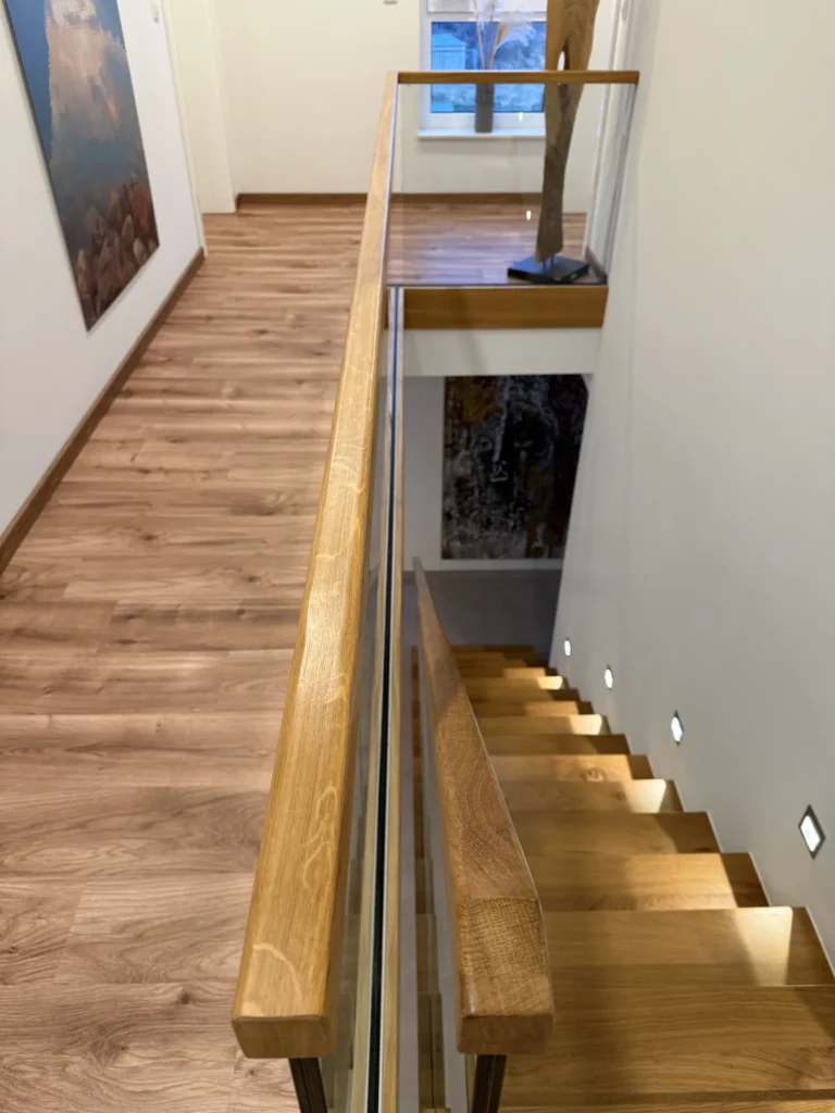 Eine moderne Innentreppe mit Holzstufen und Glasgeländer führt von einem Flur mit hellem Holzboden und weißen Wänden hinunter, der mit Kunstwerken und einem großen Fenster am Ende ausgestattet ist.