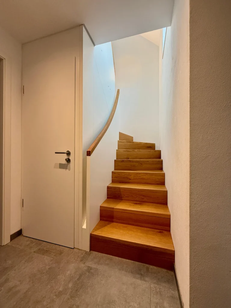 Eine moderne, schmale Holztreppe mit einem geschwungenen Handlauf führt neben einer geschlossenen weißen Tür in ein minimalistisches Interieur mit weißen Wänden und grauem Fliesenboden. Natürliches Licht erhellt den Raum.