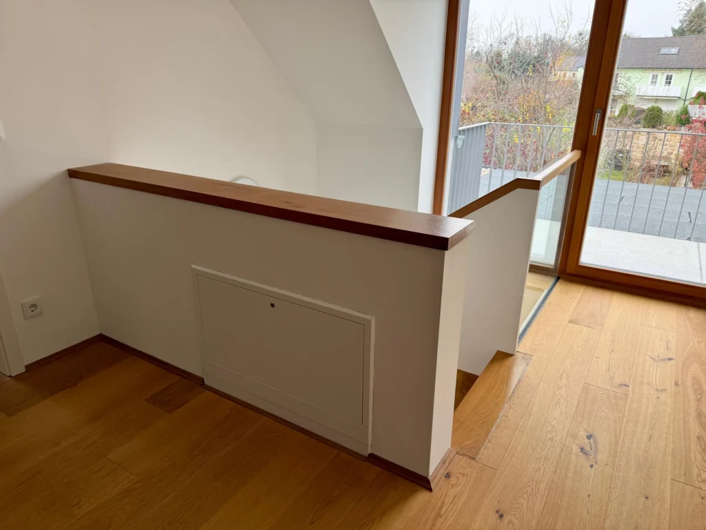 Ein kleiner Treppenabsatz am oberen Ende einer Holztreppe mit weißem Halbmauergeländer, hölzernem Handlauf und einer Tür, die auf einen Balkon mit Blick auf die Häuser und Bäume draußen führt.