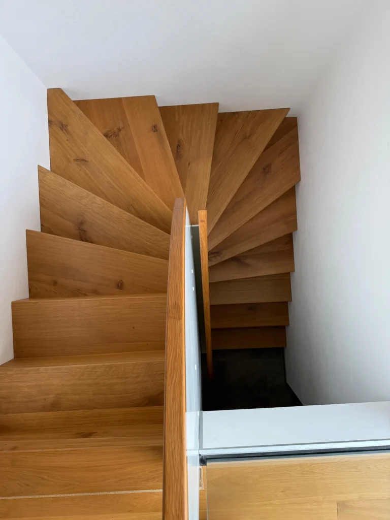 Draufsicht auf eine moderne Holztreppe mit kantigen Stufen, mit einem Geländer aus Glas und Holz auf einer Seite und weißen Wänden, die die Treppe umschließen.