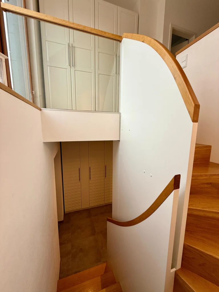 Eine moderne Innentreppe mit Holzstufen und weißem Handlauf führt zu einem Treppenabsatz mit Glas- und Holzgeländer. Sowohl in der oberen als auch in der unteren Etage sind weiße Einbauschränke zu sehen.